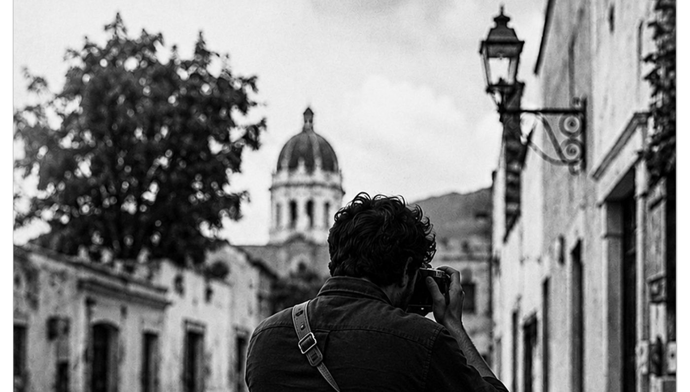 Fotógrafo de espaldas disparando una cámara de 35mm en una calle empedrada de Tequisquiapan, con la cúpula de la parroquia al fondo